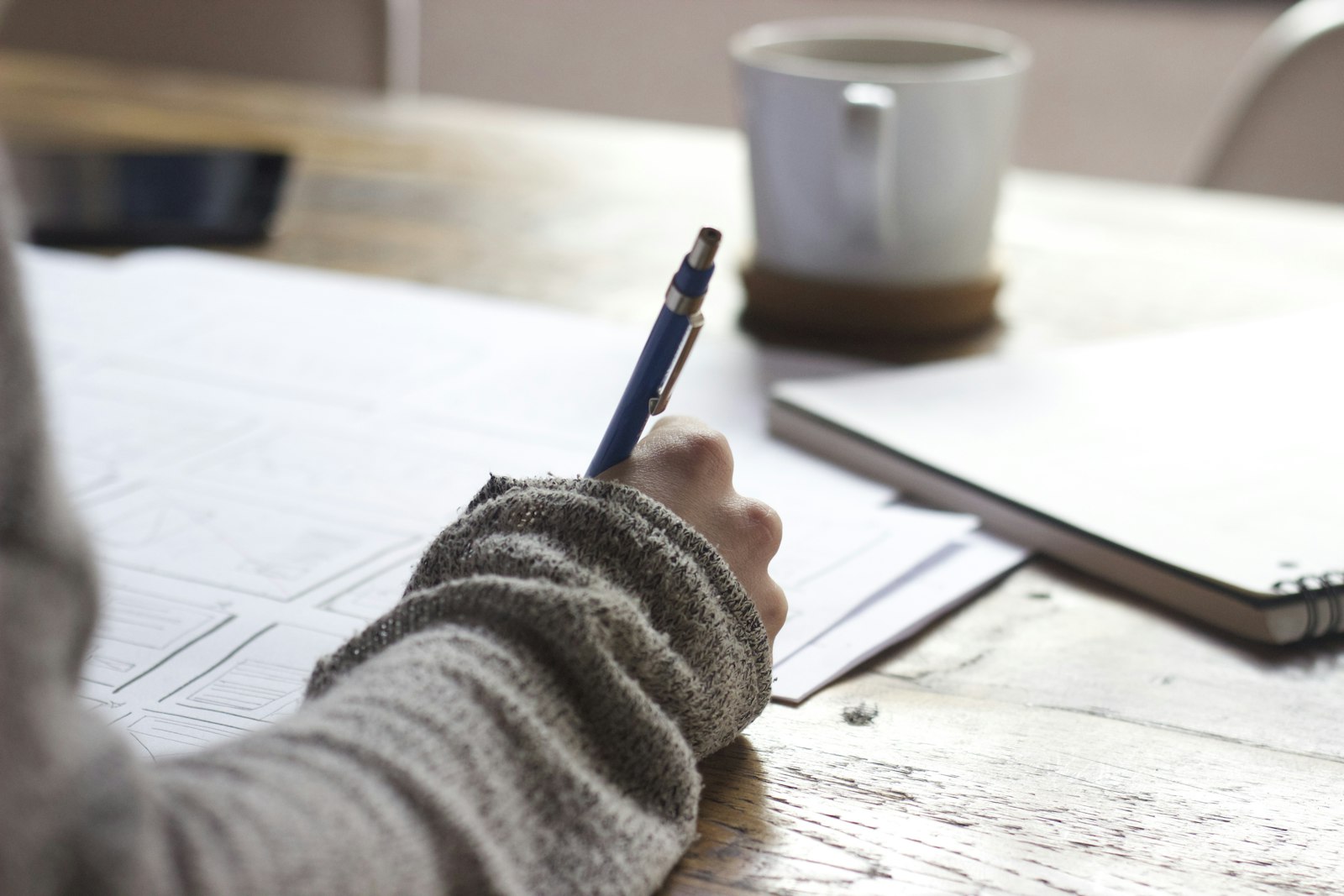 Une personne écrit dans un carnet sur une table en bois, près d'une tasse